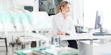Horizontal color image of female doctor sitting at her desk in dental clinic office and using computer. Female dentist wearing white lab coat and surgical mask.