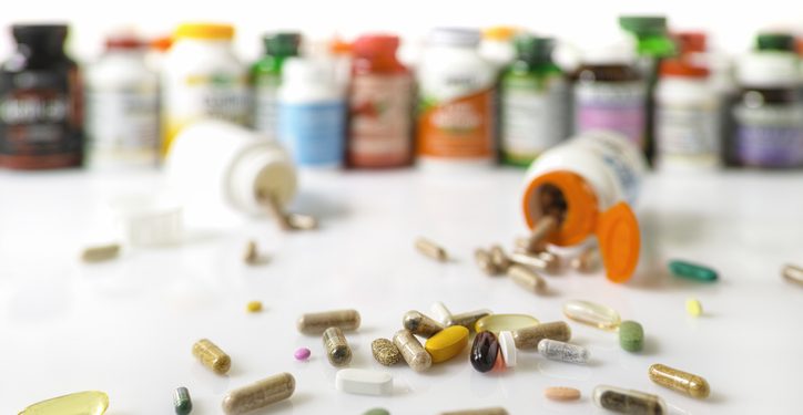 Colorful display and stack of many types of nutritional supplements and vitamin plastic bottles in an out of focus background with many supplement and vitamin capsules and pills in focus in the foreground. Scene set on a white background.