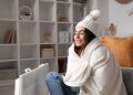 Happy young woman in winter clothes warming near radiator at home