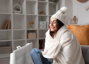 Happy young woman in winter clothes warming near radiator at home