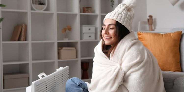 Happy young woman in winter clothes warming near radiator at home