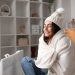Happy young woman in winter clothes warming near radiator at home